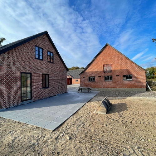 Ny terrasse og gårdsplads under etablering mellem murstensbygninger med fliser og afrettet jord.