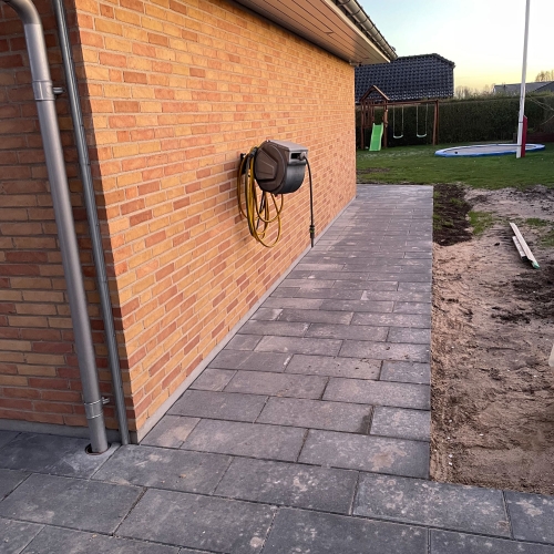 Smal flisebelægning langs murstenshus med jord klar til videre havearbejde ved siden af.