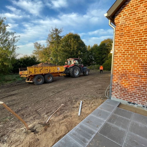 Anlægsarbejde ved murstensbygning med traktor, vogn og nylagt fliseareal.